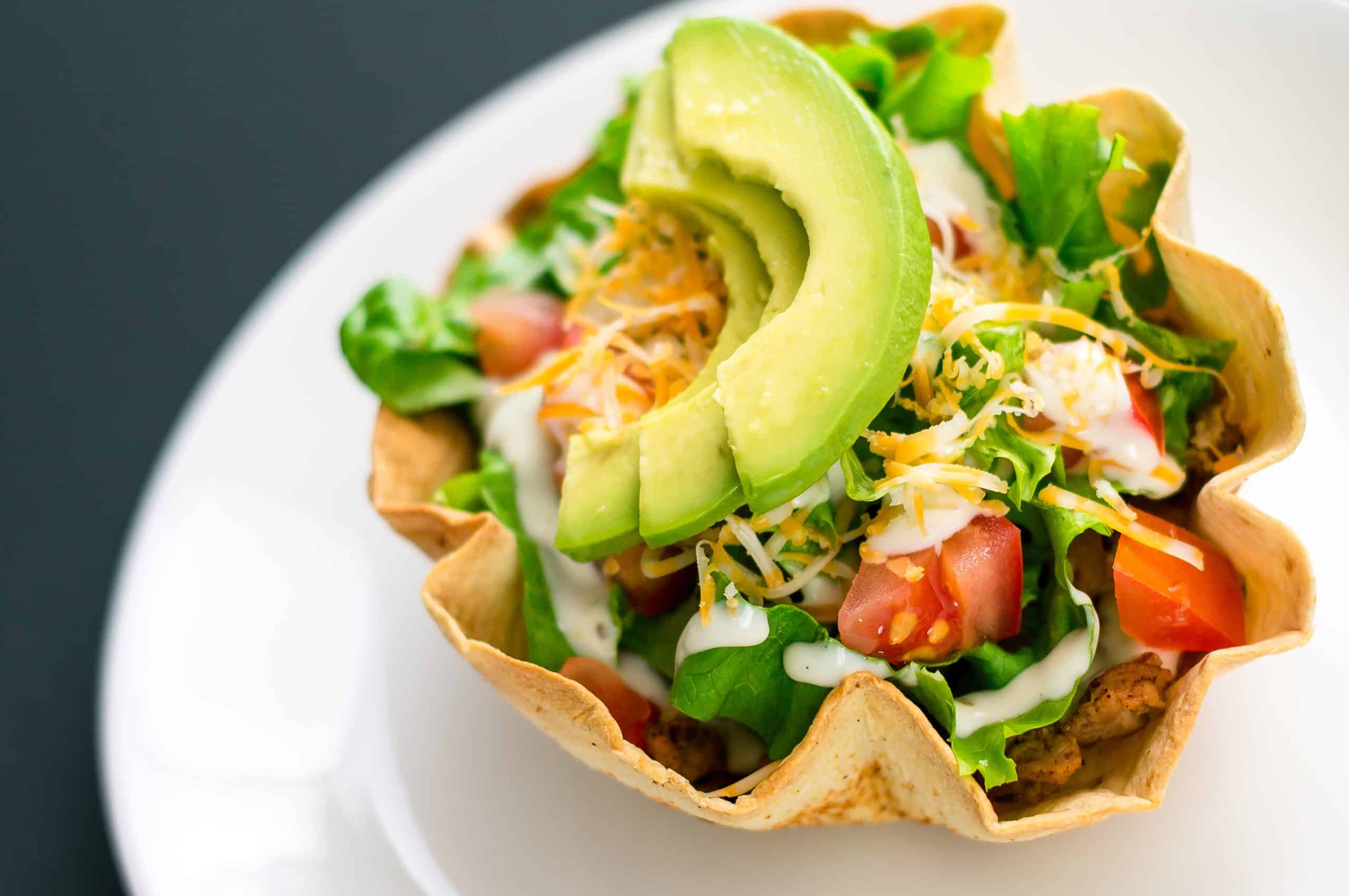 A taco salad in tortilla bowl is a fun and colorful way to eat mexican food. Made with fresh ingredients such as avocado, tomatoes, green salad, cheese and delicious greek yoghurt.